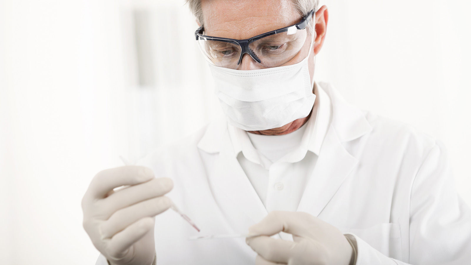Mann in weißem Kittel, mit Mundschutz und Schutzbrille nimmt eine Probe mit einer Pipette.